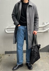 Manteau en laine grise sur un pull noir en maille, jean denim bleu et mocassins noirs. Sac fourre-tout en tissu noir avec des accents de ruban. Textures lisses mises en avant.