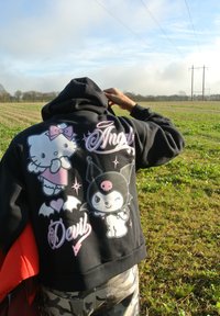 Μαύρη φούτερ με μπροστινές ροζ και λευκές καρτούν γραφικές απεικονίσεις της Hello Kitty και ενός χαρακτήρα με καπέλο νυχτερίδας, με κείμενο "Άγγελος" και "Διάβολος".