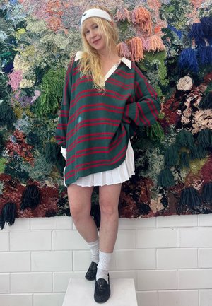 Jersey de lana verde con rayas rojas y un cuello blanco, acompañado de una falda blanca con pliegues. Se llevan zapatos negros y calcetines deportivos blancos.