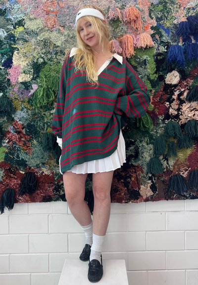 Jersey de lana verde con rayas rojas y un cuello blanco, acompañado de una falda blanca con pliegues. Se llevan zapatos negros y calcetines deportivos blancos.