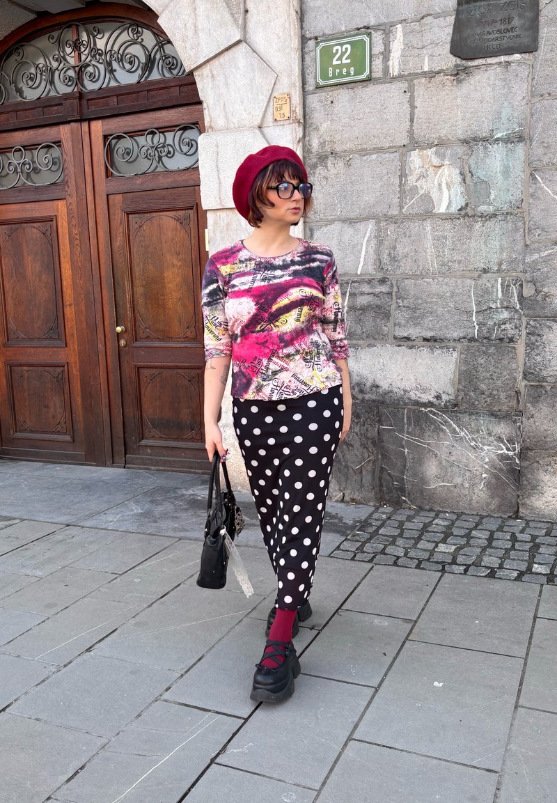 Personne portant un béret rouge, des lunettes de soleil, un haut à motifs, une jupe noire à pois blancs, des chaussettes rouges et des chaussures noires, marchant près d'un mur de pierre et d'une porte en bois.