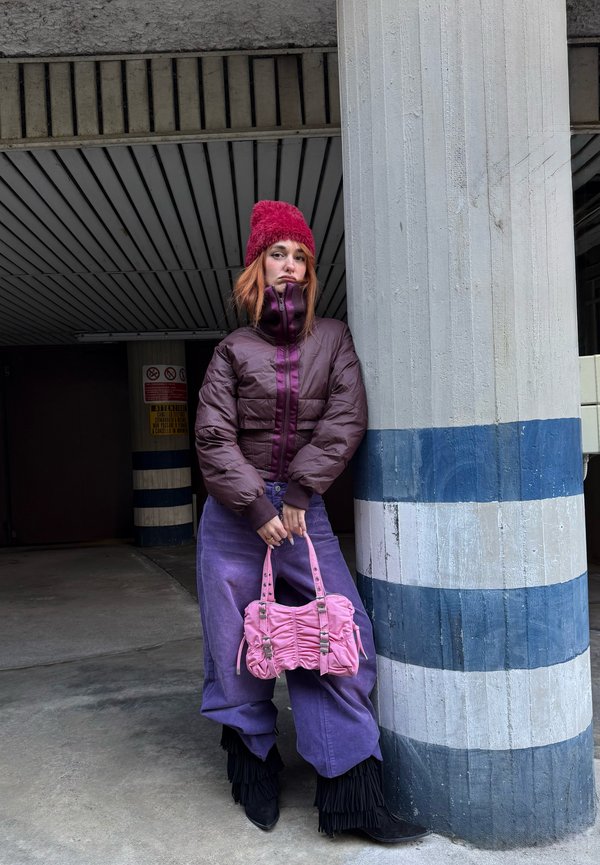 Sieviete violetā žaketē, violetās biksēs un sarkanā cepurē atbalstājas pret svītrainu kolonnas stabu, turot rokās rozā rokassomu pilsētas stāvlaukumā.