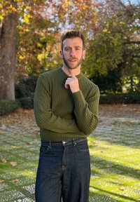 Homme aux cheveux courts et à la barbe, portant un pull vert et un jean sombre, debout en plein air en automne avec des arbres et des feuilles tombées derrière lui.