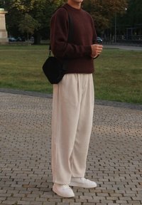 Maglione a maglia marrone, pantaloni a gamba larga a coste color crema, sneakers bianche e una borsa a tracolla nera. Ambientazione esterna con sfondo in pavimentazione di pietra.
