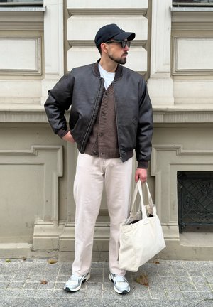 Hombre con chaqueta de cuero negra, cárdigan marrón, pantalones blancos y gorra, sosteniendo una bolsa de tela blanca, de pie en una acera de la ciudad contra una pared de piedra.