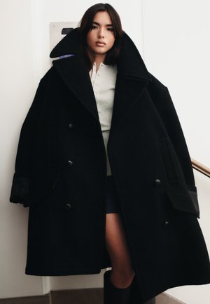 Palton oversized negru din lână, cu rever mari și design cu dublă închidere. Are buzunare laterale și o textură netedă, structurată.