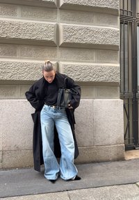 Άτομο με μαλλιά πιασμένα πίσω φοράει μαύρο μακρύ παλτό, μαύρο τοπ, φαρδιά ανοιχτό μπλε τζιν, μαύρα μυτερά παπούτσια, κρατώντας μαύρη τσάντα χειρός, στέκεται δίπλα σε πέτρινο τοίχο.