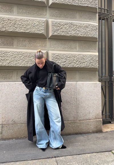 Persona con el cabello recogido, viste un abrigo largo negro, camiseta negra, jeans anchos azul claro, zapatos negros puntiagudos, sostiene un bolso negro y está junto a un muro de piedra.