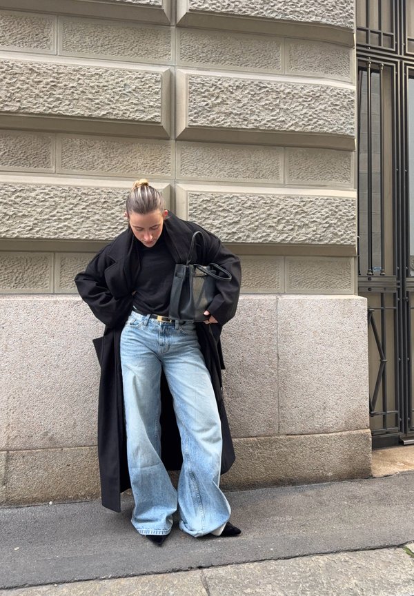 Person mit zurückgebundenem Haar trägt einen schwarzen langen Mantel, ein schwarzes Oberteil, weite hellblaue Jeans, schwarze spitze Schuhe und hält eine schwarze Handtasche, steht an einer Steinmauer.