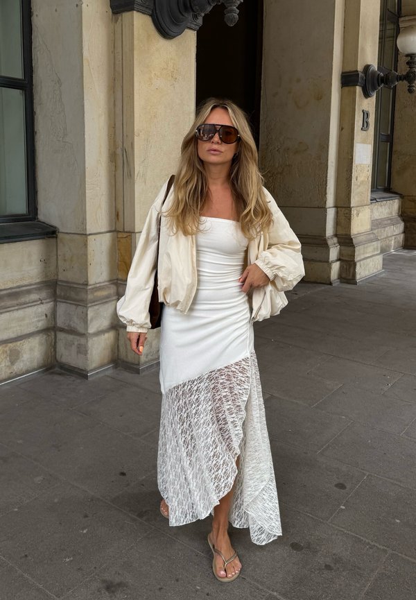 Femme aux longs cheveux blonds portant de grandes lunettes de soleil, une robe asymétrique blanche en dentelle, une veste crème et des tongs marchant sur un trottoir en pierre.