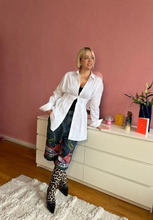 Camisa blanca de botones, jeans gráficos con diseño floral y botines de charol con estampado de leopardo. Suelo de madera, pared rosa, objetos decorativos en la cómoda.