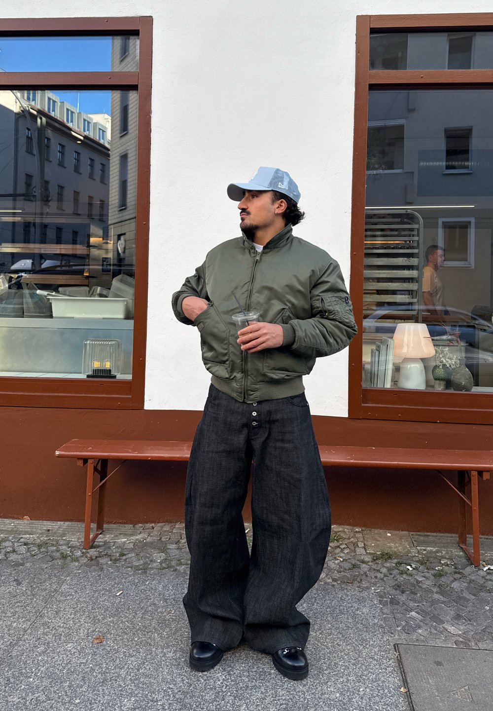 Blouson aviateur vert olive, pantalon large en denim foncé et chaussures noires. Tient un verre transparent avec une paille, debout près d'une fenêtre avec des reflets.