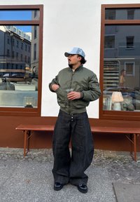 Blouson aviateur vert olive, pantalon large en denim foncé et chaussures noires. Tient un verre transparent avec une paille, debout près d'une fenêtre avec des reflets.
