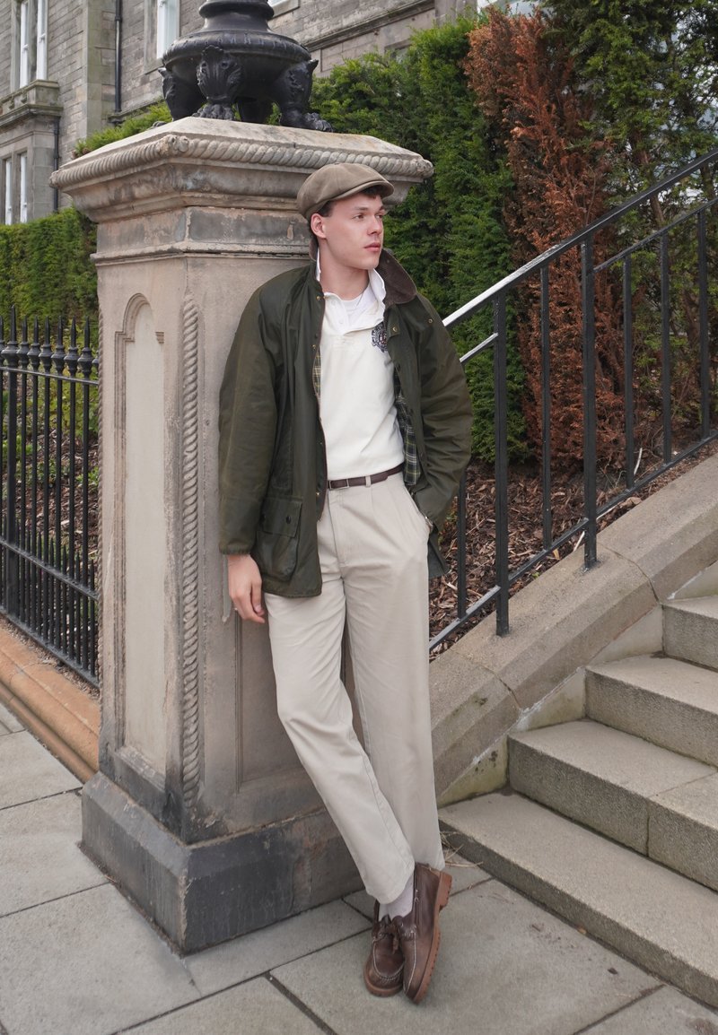 Joven con pantalones beige, zapatos marrones, chaqueta verde y gorra apoyado en una columna de piedra cerca de unas escaleras exteriores y vegetación.