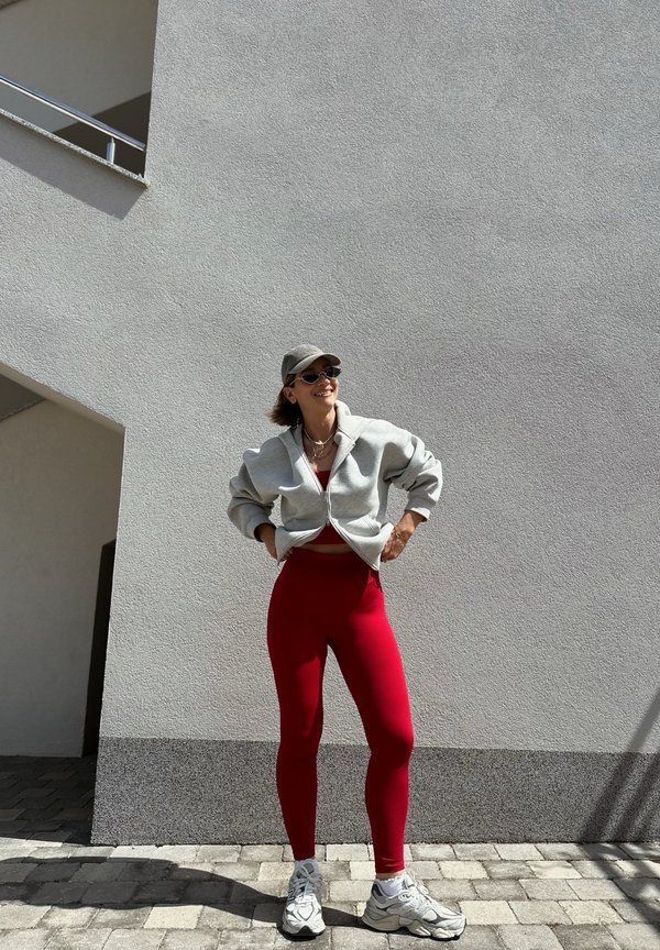 Kvinde iført en grå kasket, solbriller, en grå jakke, røde leggings og hvide sneakers står smilende ved en lysegrå væg udendørs.