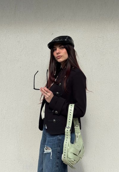 Joven mujer con chaqueta negra y gorra sostiene unas gafas de sol y un bolso verde claro contra una pared blanca texturizada.