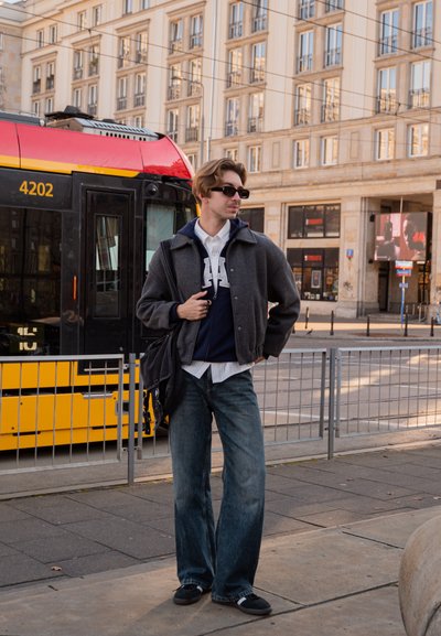 Conjunto casual que incluye una chaqueta gris oscuro, sudadera con capucha azul marino y jeans de pierna ancha. Los accesorios incluyen gafas de sol y un bolso negro. Tranvía amarillo al fondo.