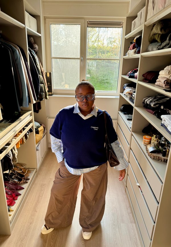 Osoba sa naočalama, tamnoplavim džemperom, prugastom košuljom i smeđim hlačama stoji u dobro organiziranom garderobnom ormaru s policama i odjećom.