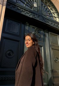 Ένα σκούρο καφέ μπουφάν με υφή που φοριέται από ένα άτομο μπροστά σε μια μεγάλη μαύρη διακοσμητική πόρτα με περίπλοκα σχέδια και λεπτο�μέρειες.