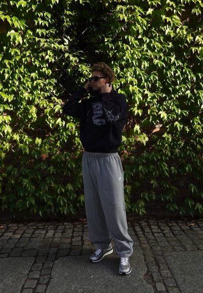 Sudadera negra con detalle gráfico, pantalones de chándal grises y zapatillas plateadas. Rodeado de hiedra verde, de pie sobre pavimento de adoquines.
