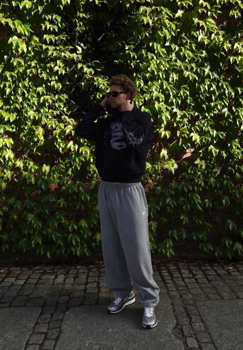 Schwarzes Sweatshirt mit grafischen Details, graue Jogginghosen und silberne Sneakers. Umgeben von grünem Efeu, stehend auf Kopfsteinpflaster.