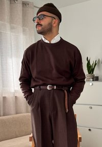 Pull en maille marron porté sur une chemise blanche à col, associé à un pantalon marron sur mesure et une ceinture en cuir marron. Lunettes noires et bonnet.