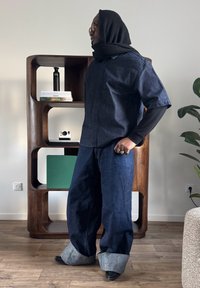 Homme portant une tenue en denim surdimensionnée avec un foulard noir, des lunettes et des bagues, debout à l'intérieur à côté d'une étagère en bois avec des livres et un appareil photo.