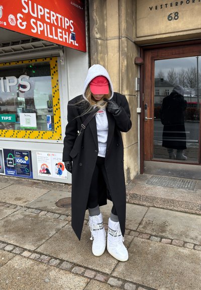 Persona con abrigo negro, sudadera con capucha gris, gorra roja y botas blancas, de pie en la acera cerca de la entrada de un edificio y una vitrina con carteles.