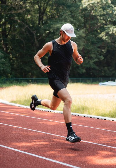 Corredor masculino con una camiseta sin mangas negra y pantalones cortos, vistiendo zapatillas deportivas negras y blancas, en una pista de atletismo roja rodeada de vegetación.