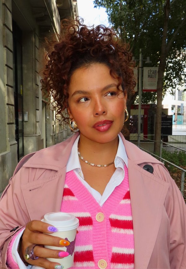 A person holds a coffee cup, wearing a pink trench coat over a striped pink sweater, white shirt, pearl necklace, and colorful manicured nails.