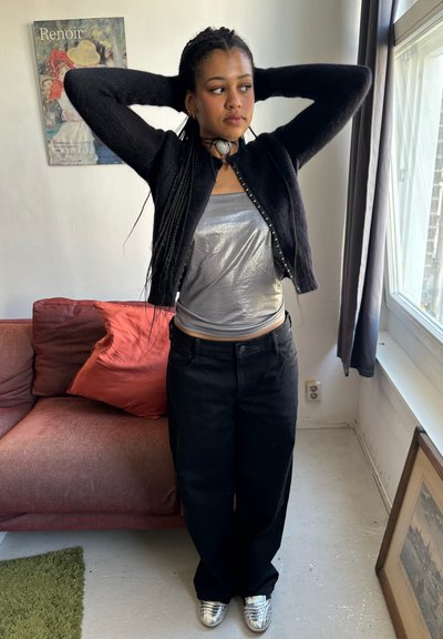 Cardigan negro de punto sobre una top de tirantes metálico plateado, combinado con pantalones anchos negros y zapatillas plateadas brillantes. Cojines rojos y marrones al fondo.