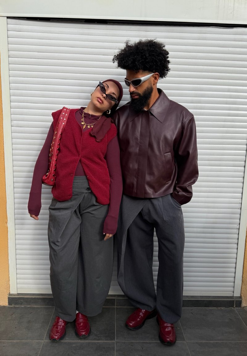 Deux personnes vêtues de tenues bordeaux et grises se tiennent côte à côte ; la femme appuie sa tête sur l'épaule de l'homme contre un volet blanc.