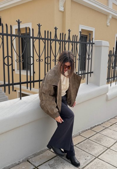 Mujer con gafas de sol que lleva una chaqueta marrón, una camiseta beige y pantalones negros, sentada sobre un muro blanco junto a una reja de hierro negra en un camino de baldosas.