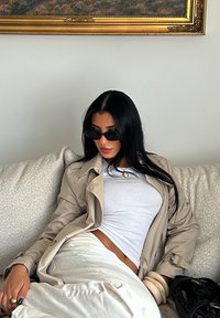 Beige trenchcoat, hvit tettsittende topp, hvit skjørt. Svart solbriller, gullsmykker, chunky armbånd. Myk, teksturert sofa, nøytral bakgrunn.