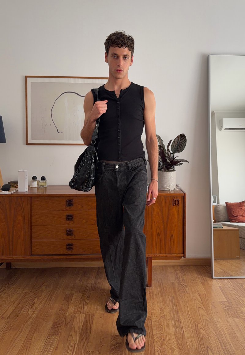 Homme en haut noir sans manches et pantalon noir ample marche à l’intérieur, portant un sac noir ; commode en bois, plante et grand miroir en arrière-plan.