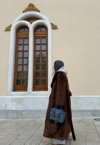 Personne en long manteau marron et sweat à capuche gris portant un sac à main bleu marchant devant un mur beige avec de grandes fenêtres en bois cintrées.