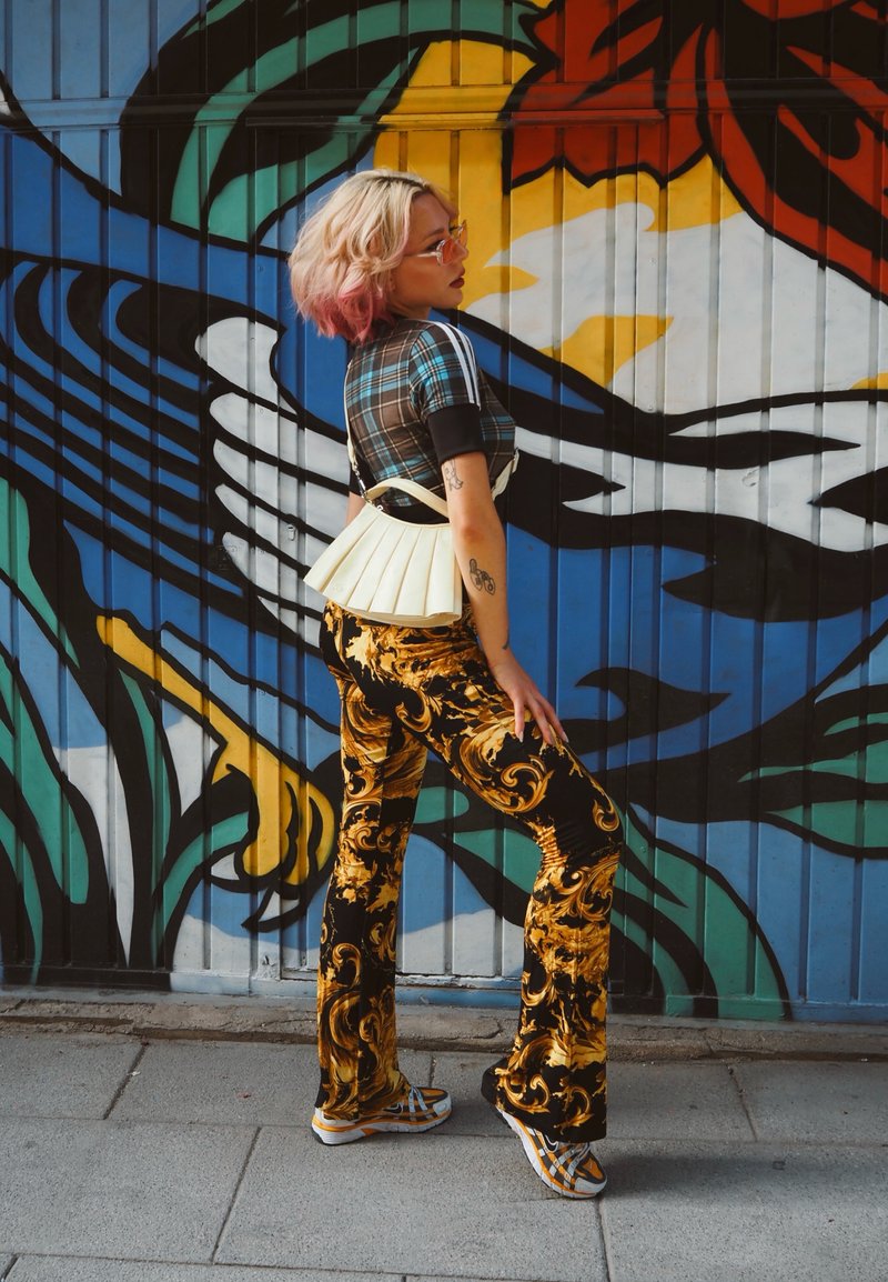 Femme aux cheveux blonds avec des pointes roses, portant un pantalon à motifs et un haut à carreaux, posant de profil devant une fresque murale abstraite colorée.