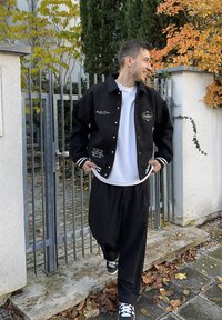 Must ülikoolijakk valgete triipude ja tikitud kirjaga, lõdva lõikega. Sobitatud mustade lahtiste pükste ja mustade tossudega.