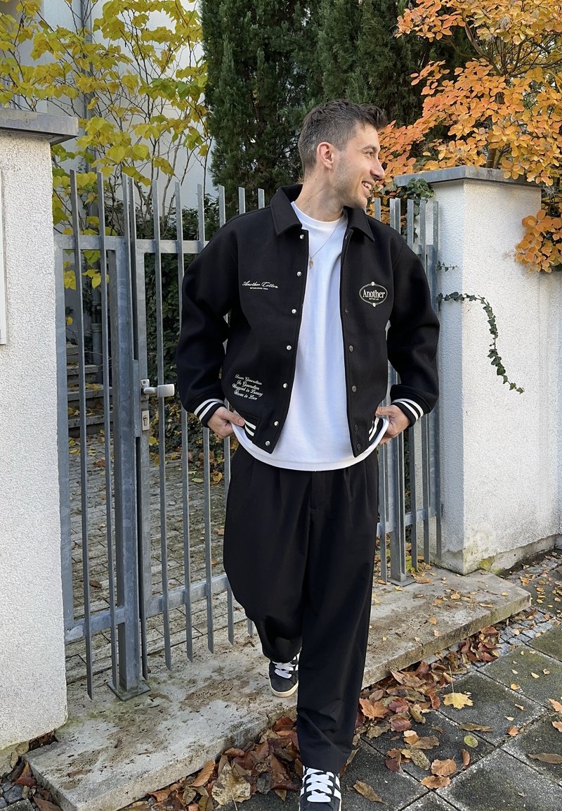 Svart varsityjacka med vita ränder, broderad text och avslappnad passform. Kombinerad med svarta lösa byxor och svarta sneakers.