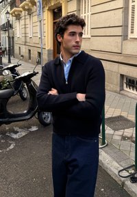 Jeune homme aux cheveux bouclés portant un pull noir zippé et un jean bleu, debout les bras croisés sur un trottoir en ville près de scooters stationnés.