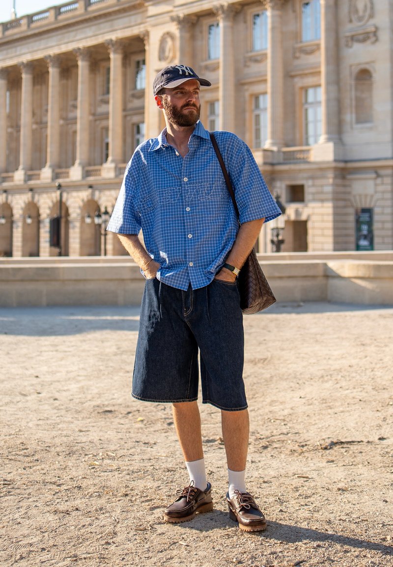 Homme avec barbe portant une chemise à carreaux bleue, un short en jean, des chaussettes blanches, des chaussures marron et une casquette foncée se tient dehors devant un bâtiment classique.