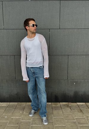 Camisa blanca de manga larga con mangas transparentes, combinada con unos jeans holgados azul claro y zapatillas plateadas. Frente a una pared gris oscura.
