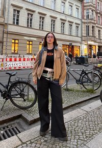 Veste en velours côtelé beige, t-shirt noir avec logo, ceinture à motifs, pantalons larges noirs, bottes noires. Décor urbain avec des vélos et des bâtiments.