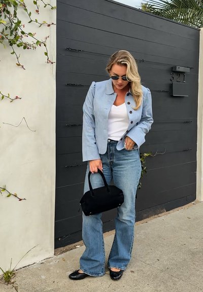 Mujer con cabello largo y rubio que lleva gafas de sol, blazer azul claro, camiseta blanca, vaqueros azules, zapatos planos negros, sujetando un bolso negro junto a una puerta oscura.