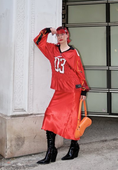 Jersey rojo de gran tamaño con acentos negros y el número 03, combinado con una falda de satén rojo brillante y botas negras hasta la rodilla; sosteniendo un bolso naranja.