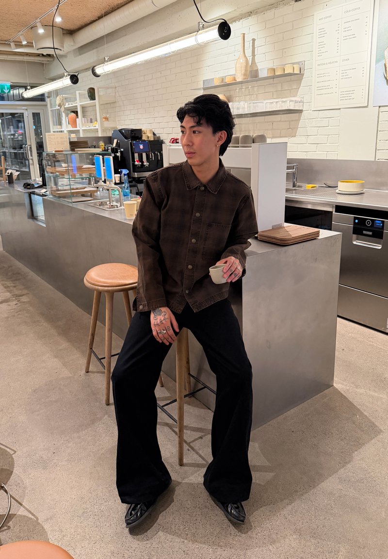 Personne avec des tatouages sur les mains assise sur un tabouret dans un café moderne, tenant une petite tasse et regardant sur le côté.