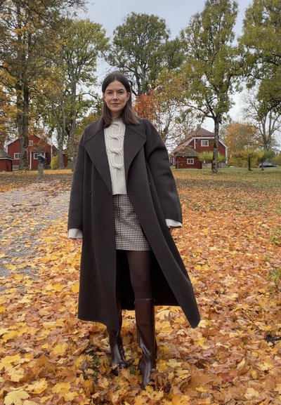 Abrigo largo marrón sobre un suéter de punto de cable color crema, combinado con una falda corta de cuadros y botas marrones hasta la rodilla, sobre un suelo cubierto de hojas.