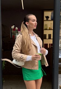 Bež kraadine jakk nööbiga, valge ribadega topp, roheline mini seelik, hoides rohelist jooki klaasis, seistes klaasuste lähedal.