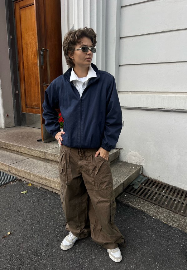 Persona con gafas de sol, chaqueta azul marino, camisa blanca y pantalones cargo marrones y holgados, de pie en la acera cerca de unos escalones de piedra y una puerta de madera.