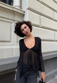 Nero maglietta a maniche lunghe con scollo a V profondo e dettaglio annodato nella parte anteriore, sovrapposta a un tessuto trasparente e fluido. Indossata con jeans scuri a vita alta.